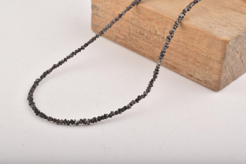 Puede incluir: Un collar de cuentas negras compuesto por peque&ntilde;as cuentas de forma irregular. El collar est&aacute; colocado sobre un bloque de madera, sobre un fondo blanco. Las cuentas parecen ser de color gris oscuro.