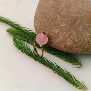 May include: A pink carved gemstone ring with a small red gemstone accent. The ring is set in gold and is displayed on a rock and green foliage.