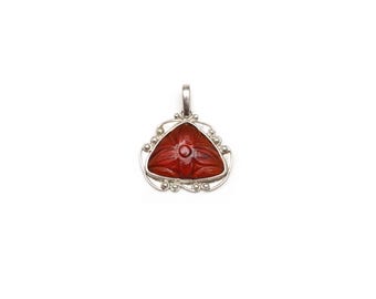 May include: A triangular pendant featuring a deep red carved gemstone with a floral motif. The stone is set within a silver frame that displays intricate scrollwork, small spherical accents, and a simple bail at the top for attachment.