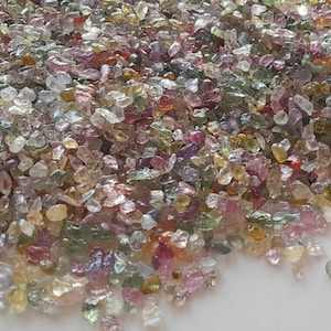 May include: A pile of small, colorful gemstones in shades of pink, green, yellow, and purple. The gemstones are irregularly shaped and appear to be raw or uncut. The stones are scattered across a white surface.