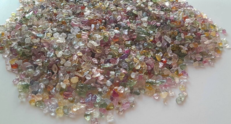 May include: A pile of small, colorful gemstones in shades of pink, green, yellow, and purple. The gemstones are irregularly shaped and appear to be raw or uncut. The stones are scattered across a white surface.