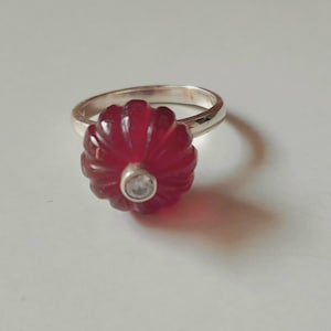 May include: A silver ring with a red gemstone set in a flower shape. The gemstone has a small clear stone in the center.