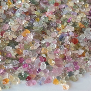 May include: A close-up view of a pile of small, colorful gemstones. The gems are a mix of colors, including pink, yellow, green, and clear. The stones are irregularly shaped and appear to be raw or uncut. The overall effect is one of natural beauty and variety.