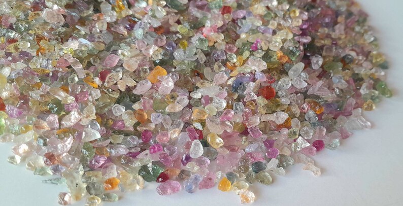 May include: A close-up view of a pile of small, colorful gemstones. The gems are a mix of colors, including pink, yellow, green, and clear. The stones are irregularly shaped and appear to be raw or uncut. The overall effect is one of natural beauty and variety.