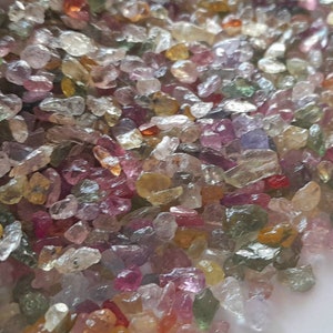 May include: A close-up view of a pile of small, colorful gemstones. The gems are a mix of colors, including pink, green, yellow, and clear. They are irregularly shaped and appear to be raw or uncut. The gemstones are scattered across a white surface.