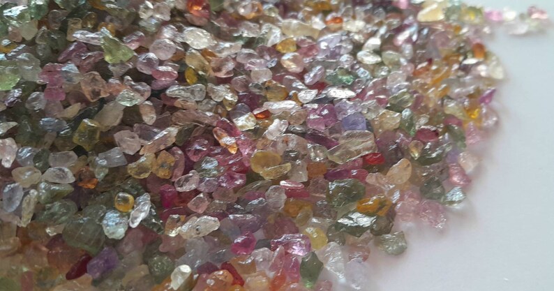 May include: A close-up view of a pile of small, colorful gemstones. The gems are a mix of colors, including pink, green, yellow, and clear. They are irregularly shaped and appear to be raw or uncut. The gemstones are scattered across a white surface.