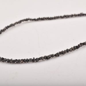 Puede incluir: Un collar compuesto por peque&ntilde;as cuentas de color gris oscuro, de forma irregular. Las cuentas est&aacute;n ensartadas, formando una hebra continua. El collar se muestra sobre un fondo blanco, resaltando la textura y el color de las cuentas.