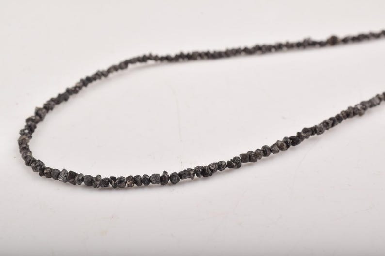 Puede incluir: Un collar compuesto por peque&ntilde;as cuentas de color gris oscuro, de forma irregular. Las cuentas est&aacute;n ensartadas, formando una hebra continua. El collar se muestra sobre un fondo blanco, resaltando la textura y el color de las cuentas.