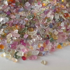 May include: A pile of small, colorful gemstones in various shades of pink, purple, yellow, green, and orange. The gemstones are irregularly shaped and appear to be raw or uncut, with a slightly translucent quality.