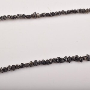 Puede incluir: Dos hilos de cuentas de diamantes en bruto de color gris oscuro. Las cuentas tienen forma irregular y est&aacute;n ensartadas, creando una est&eacute;tica cruda y natural. El fondo es una superficie blanca lisa.