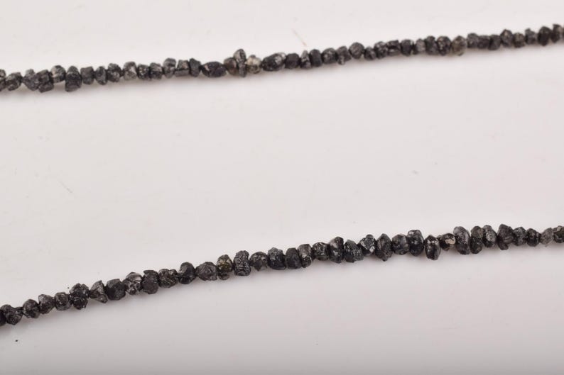 Puede incluir: Dos hilos de cuentas de diamantes en bruto de color gris oscuro. Las cuentas tienen forma irregular y est&aacute;n ensartadas, creando una est&eacute;tica cruda y natural. El fondo es una superficie blanca lisa.