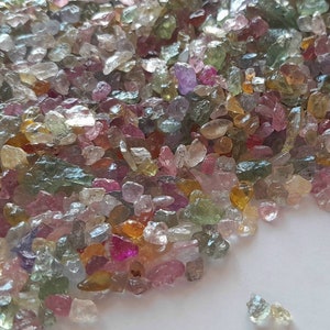 May include: A collection of small, rough-cut gemstones in various colors, including pink, green, yellow, and clear. The gemstones are scattered across a white surface, creating a colorful and textured display. The stones appear to be raw and natural.