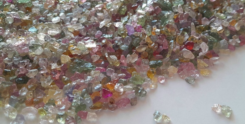 May include: A collection of small, rough-cut gemstones in various colors, including pink, green, yellow, and clear. The gemstones are scattered across a white surface, creating a colorful and textured display. The stones appear to be raw and natural.