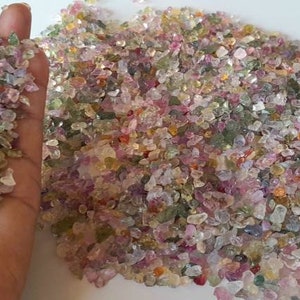 May include: A collection of small, colorful gemstones in shades of pink, green, and clear. The gemstones are irregularly shaped and appear to be tumbled. A handful of the gemstones are held in a hand.