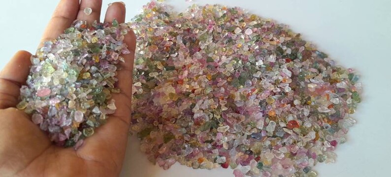 May include: A collection of small, colorful gemstones in shades of pink, green, and clear. The gemstones are irregularly shaped and appear to be tumbled. A handful of the gemstones are held in a hand.