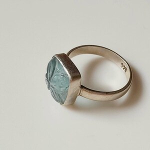 May include: A silver ring with a square, light blue gemstone. The gemstone has a carved floral design. The ring band is thin and has the number "925" stamped on it. The ring is set against a white background.