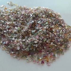 May include: A pile of small, irregularly shaped gemstones in various colors, including pink, green, yellow, and clear. The gemstones are scattered across a white surface, creating a sparkling effect.