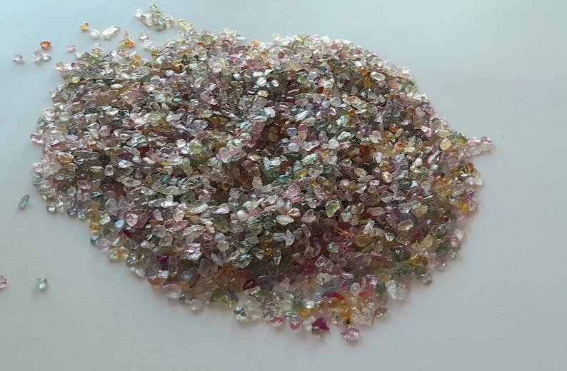 May include: A pile of small, irregularly shaped gemstones in various colors, including pink, green, yellow, and clear. The gemstones are scattered across a white surface, creating a sparkling effect.