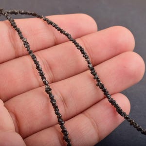 Puede incluir: Un hilo de peque&ntilde;as cuentas de diamante en bruto de color gris oscuro. Las cuentas est&aacute;n ensartadas, formando un collar o una pulsera. Las cuentas se sostienen en la mano de una persona sobre un fondo oscuro.