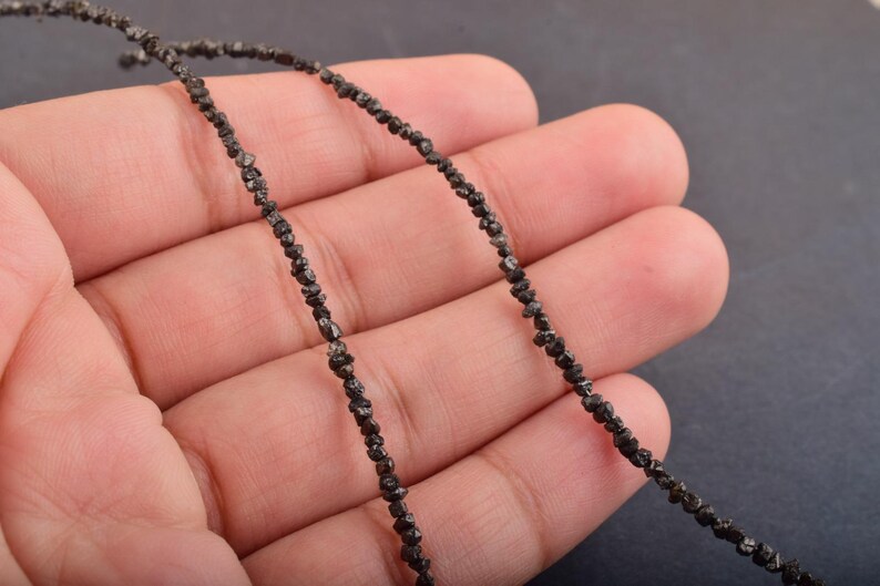 Puede incluir: Un hilo de peque&ntilde;as cuentas de diamante en bruto de color gris oscuro. Las cuentas est&aacute;n ensartadas, formando un collar o una pulsera. Las cuentas se sostienen en la mano de una persona sobre un fondo oscuro.