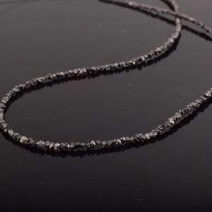 Puede incluir: Un collar gris oscuro compuesto por peque&ntilde;as cuentas de forma irregular. Las cuentas est&aacute;n ensartadas para formar una hebra continua. El collar se muestra sobre una superficie oscura y reflectante, resaltando su textura y color.