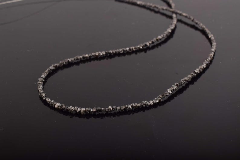 Puede incluir: Un collar gris oscuro compuesto por peque&ntilde;as cuentas de forma irregular. Las cuentas est&aacute;n ensartadas para formar una hebra continua. El collar se muestra sobre una superficie oscura y reflectante, resaltando su textura y color.