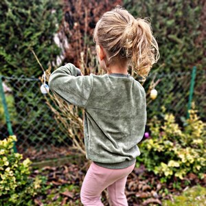 Könnte beinhalten: Eine junge Person trägt ein grünes Sweatshirt und eine rosa Hose und steht in einem grasbewachsenen Bereich. Sie trägt bunte Turnschuhe und hat ihre Haare zu einem Pferdeschwanz gebunden.