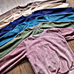 Könnte beinhalten: Ein Stapel von zehn weichen, plüschigen Sweatshirts in verschiedenen Farben, darunter Braun, Beige, Blau, Grün und Pink. Die Sweatshirts sind gefaltet und in Form eines Fächer angeordnet, wobei das rosafarbene Sweatshirt oben liegt.