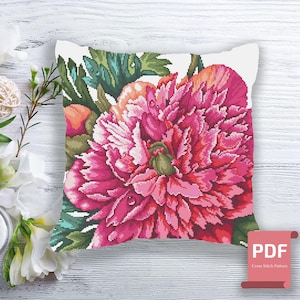 Könnte beinhalten: Ein weißes Kissen mit einem Kreuzstichmuster einer rosa Pfingstrose mit grünen Blättern. Das Kissen liegt auf einer weißen Holzoberfläche. PDF-Kreuzstichmuster.