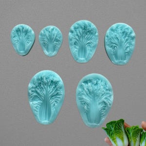 Peut inclure: Collection de six moules en silicone bleu clair en forme de feuilles de chou napa. Les moules sont de tailles variables et sont disposés sur un fond gris. Une main tient trois feuilles de chou napa miniatures dans le coin inférieur droit.