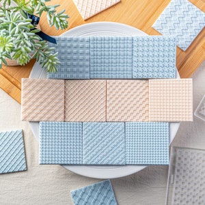 Peut inclure: Un ensemble de 12 moules en silicone carrés avec différents motifs en relief. Les moules sont disposés en cercle sur une assiette blanche. Les motifs comprennent un motif gaufré, un motif en losange, un motif à carreaux et un motif en chevrons. Les moules sont bleu clair et beige.