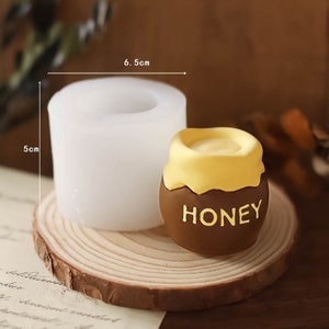Puede incluir: Un molde de silicona blanco para hacer una vela con forma de tarro de miel. El molde mide 6,5 cm de ancho y 5 cm de alto. El molde está al lado de una vela con forma de tarro de miel marrón con una parte superior de cera amarilla y la palabra "HONEY" escrita en la parte delantera.