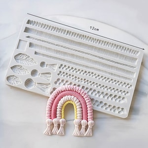 Könnte beinhalten: Eine Silikonform zum Herstellen von Kuchendekorationen. Die Form bietet eine Vielzahl von Designs, darunter Zöpfe, Federn und Perlen. Die Form ist 12 cm lang. Eine rosafarbene, gelbe und weiße Regenbogen-Kuchendeko ist neben der Form abgebildet.