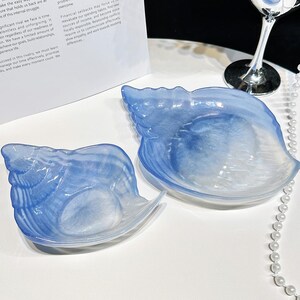 May include: Two seashell-shaped dishes in blue and white. The dishes have a translucent appearance, with a swirling pattern of colour. They are placed on a white surface, suggesting a decorative or serving purpose.