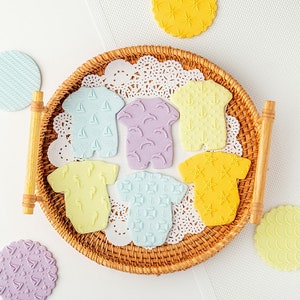 May include: A set of seven baby onesie-shaped cookie cutters, each with a different pattern. The patterns include sailboats, dolphins, anchors, seahorses, stars, and geometric designs. The cutters are made of a light blue, purple, yellow, and light green fondant.