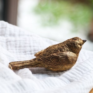 Könnte beinhalten: Detaillierte, goldfarbene Vogelfigur mit strukturierter Oberfläche. Der Vogel sitzt auf einem weißen, strukturierten Tuch. Die Figur hat einen kleinen, dunklen Schnabel und besteht wahrscheinlich aus einem haltbaren Material, das für die Inneneinrichtung geeignet ist.