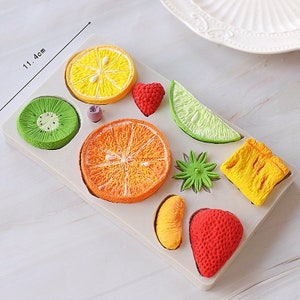 May include: A silicone mold for baking fruit-shaped cakes. The mold has 10 cavities, including a kiwi, lemon, orange, lime, strawberry, and a flower. The mold is 11.4 cm long.
