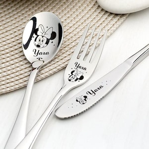 May include: A set of three silver utensils with a black and white cartoon character design. The utensils are engraved with the name "Yara".