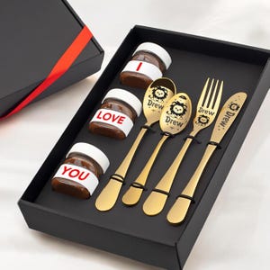 May include: A black gift box with a red ribbon contains four small jars of chocolate spread labeled "I", "LOVE", "YOU" and a gold spoon set with the name "Drew" engraved on each piece.