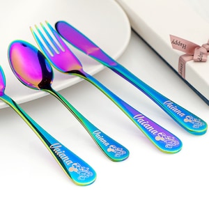 May include: Set of four rainbow iridescent stainless steel children's cutlery with the name "Vaiana" engraved on each piece. The cutlery includes a spoon, fork, and knife.