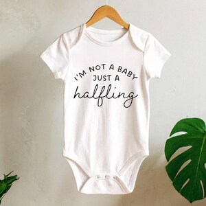 May include: White baby onesie with the text "I'M NOT A BABY JUST A halfling" in black script. The onesie is hanging on a wooden hanger against a neutral background with plants.