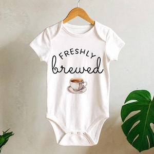 May include: White baby onesie with the words "freshly brewed" in a script font, along with a coffee cup graphic. The onesie hangs on a wooden hanger against a neutral background, with plants in the foreground.