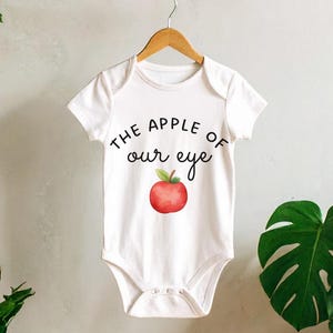 May include: A white short-sleeved baby bodysuit featuring the black text THE APPLE OF our eye and a watercolor-style red apple illustration with a green leaf centered on the front. The garment has a round neckline and snap closures at the bottom.