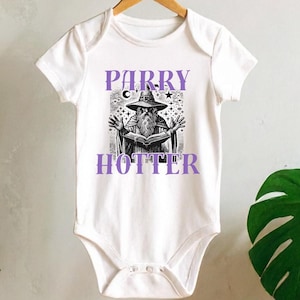 May include: White baby onesie with a purple graphic of a wizard reading a book, with the text "PARRY HOTTER". The onesie is hanging on a wooden hanger against a neutral background, with plants in the foreground.