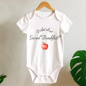 May include: White baby onesie with the text "What about Second Breakfast?" in black script and a red apple illustration. The onesie is hanging on a wooden hanger against a neutral background with plants.