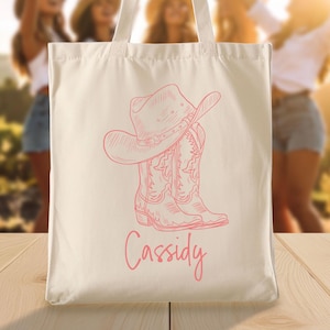 May include: A cream-colored canvas tote bag featuring a pink line-art illustration of a cowboy hat resting on a pair of cowboy boots. Below the illustration, the name Cassidy is printed in a pink cursive script font on the front of the bag.