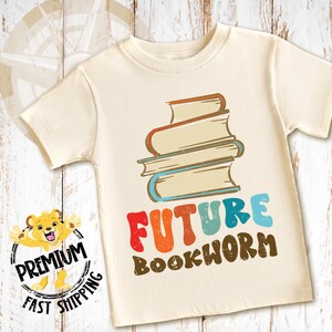 Pu&ograve; includere: Una maglietta color crema con una pila di libri e il testo "FUTURE BOOKWORM" in lettere retr&ograve; colorate.