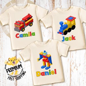 May include: Three cream-colored t-shirts with colorful graphics of toy vehicles and robots. Each shirt features a different design: a red fire truck, a yellow train, and a robot. Names "Camila", "Jack", and "Daniel" are printed below the graphics.
