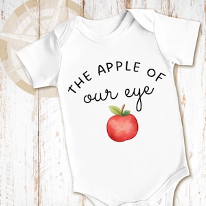 May include: A white short-sleeved baby bodysuit featuring the black printed text THE APPLE OF our eye and a watercolor-style illustration of a red apple with a green leaf positioned directly below the text on the front of the garment.