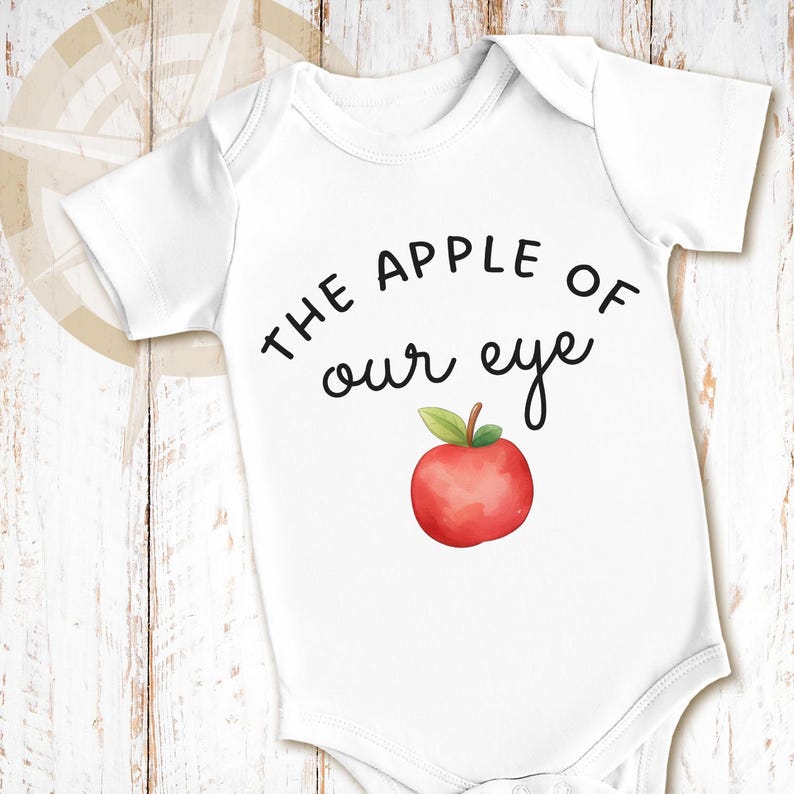 May include: A white short-sleeved baby bodysuit featuring the black printed text THE APPLE OF our eye and a watercolor-style illustration of a red apple with a green leaf positioned directly below the text on the front of the garment.